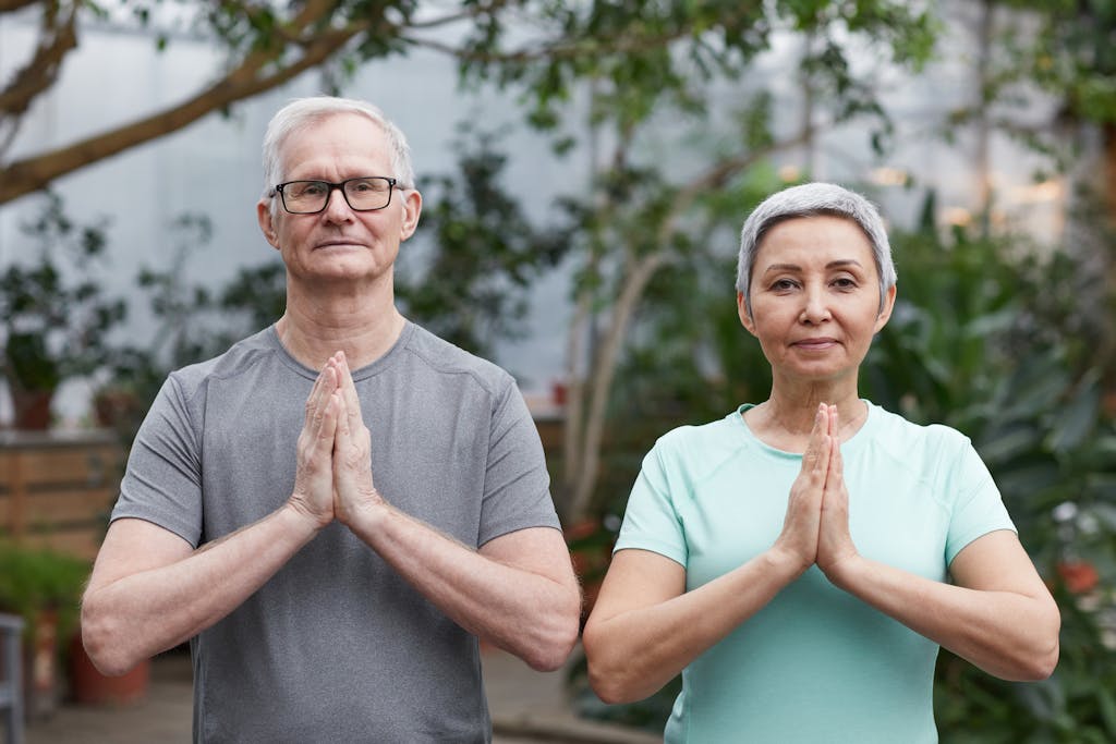 Senior couple performing yoga poses in a peaceful greenhouse setting.